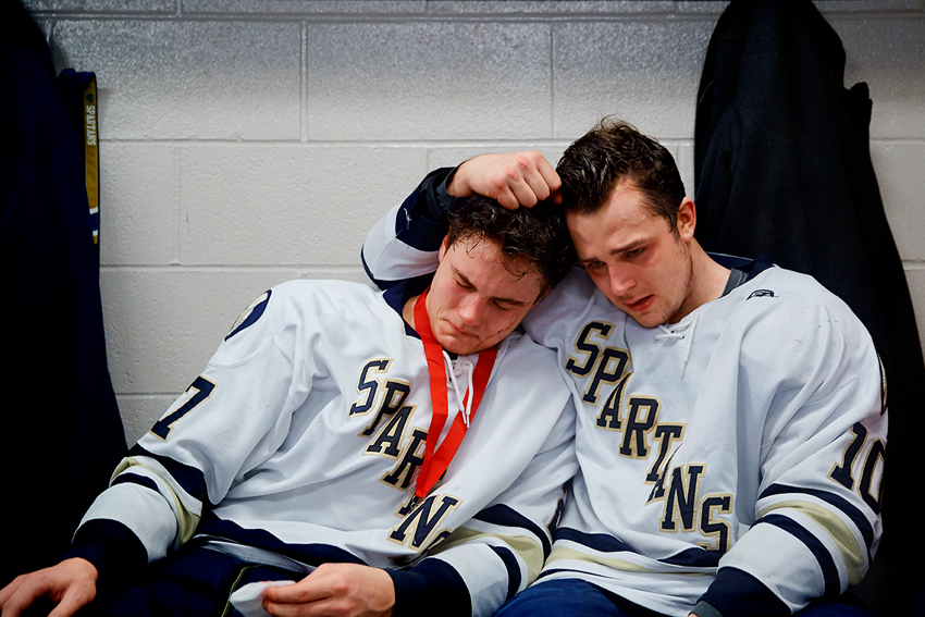 A photo by David Ellis of two high school aged ice hockey players sitting on a bench comforting each other. The boy on the left has a dried streak of blood on his face, and both boys are drenched in sweat and have emotional facial expressions.